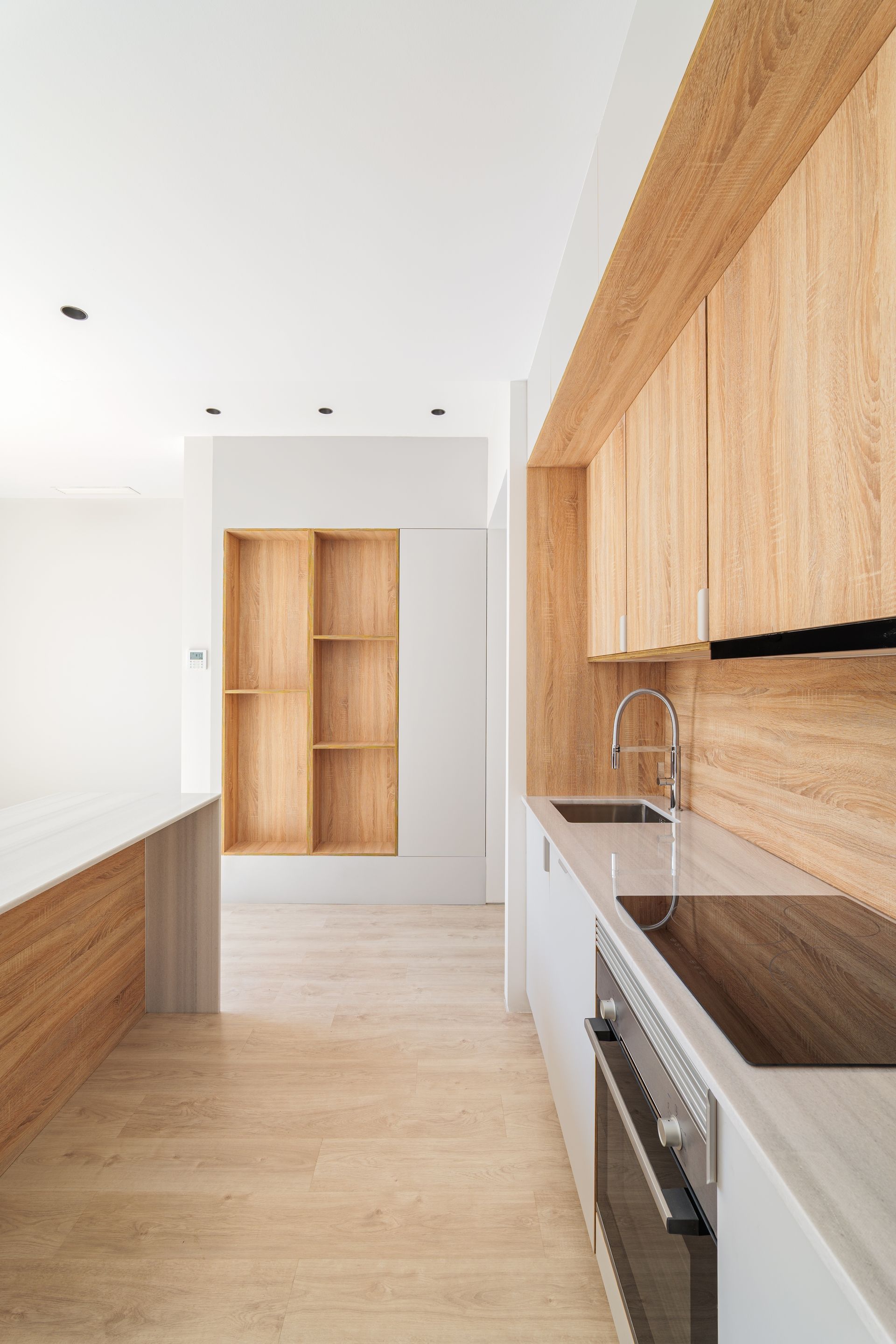 Modern kitchen with light wood cabinets, white walls, and a narrow hallway view toward built-in shelving