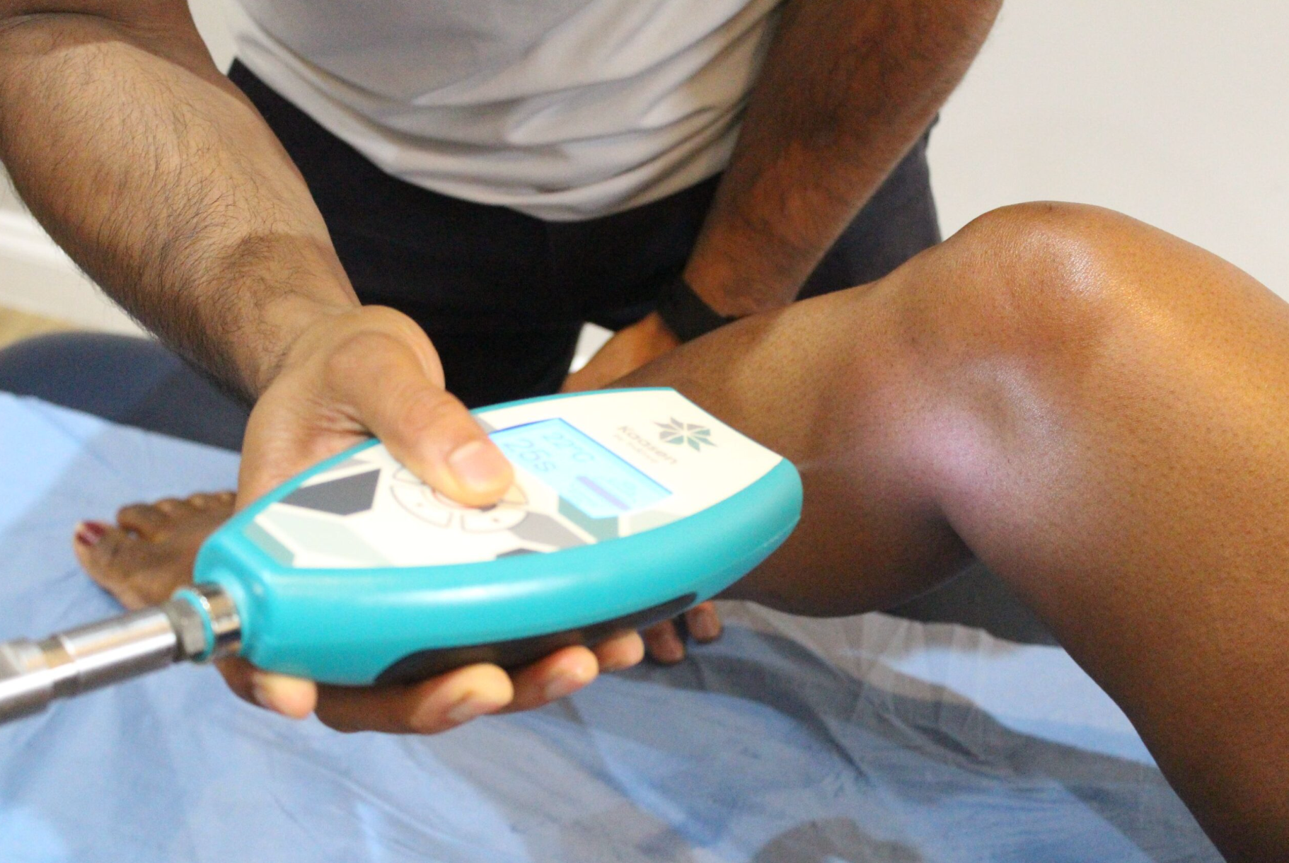 Person using a medical device on a knee. The device is teal, the leg is brown, and setting is medical.