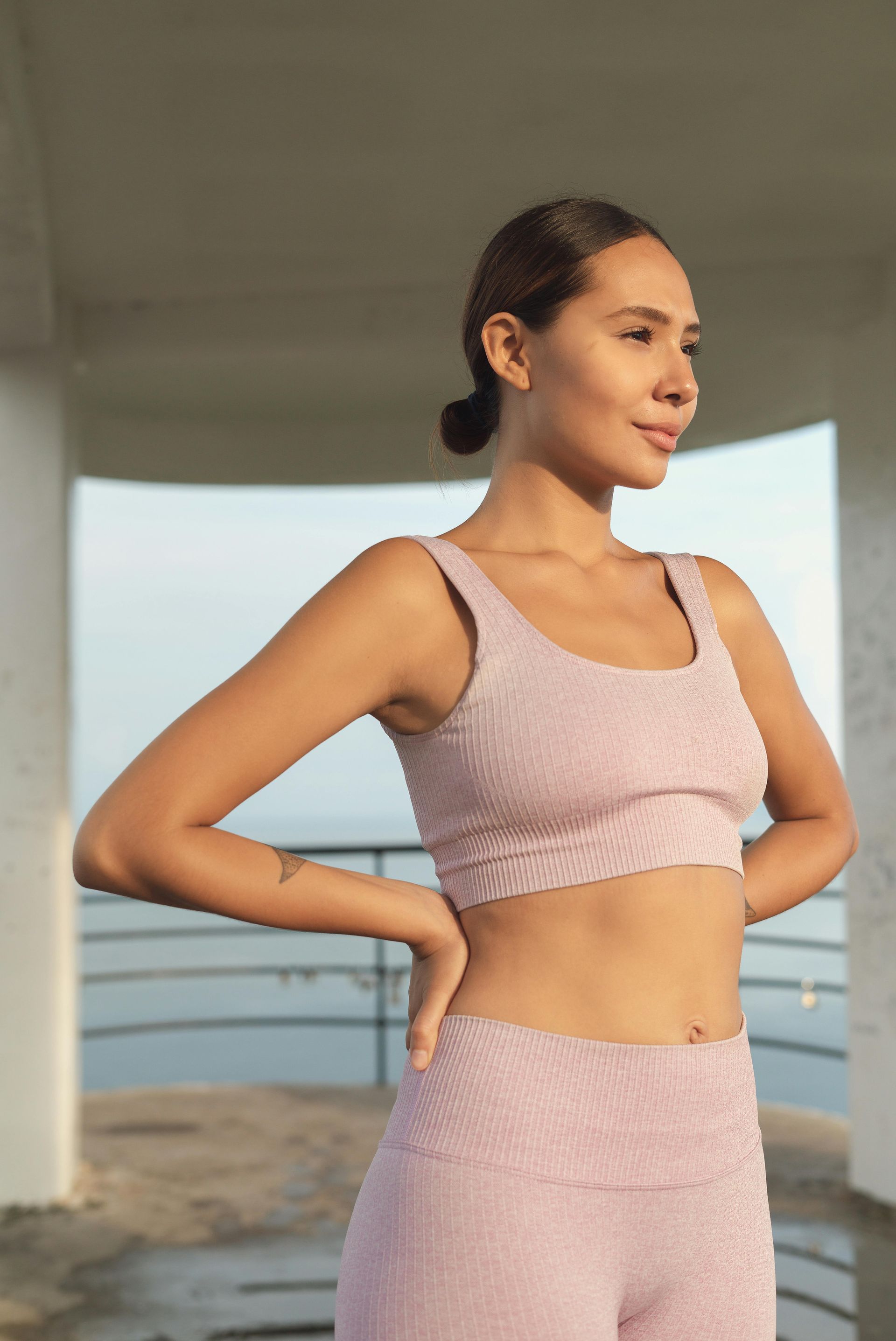 Woman in pink exercise wear, hands on hips, looking off to the side. Outdoors by a railing.