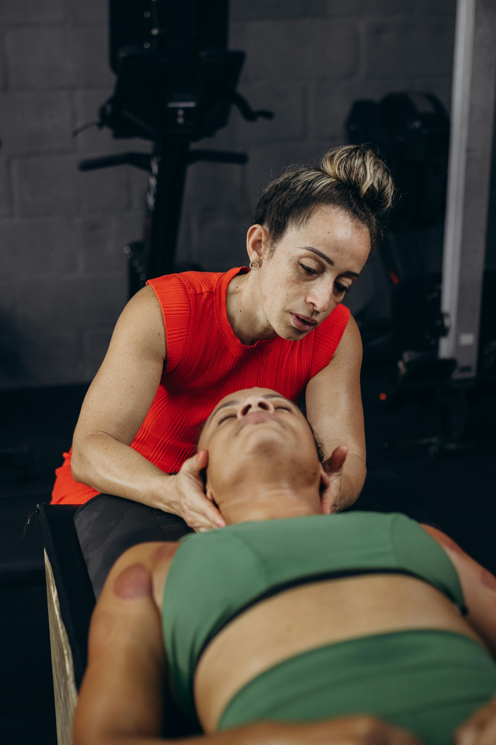 Woman in red top giving neck massage to person with cupping marks on back, gym setting.