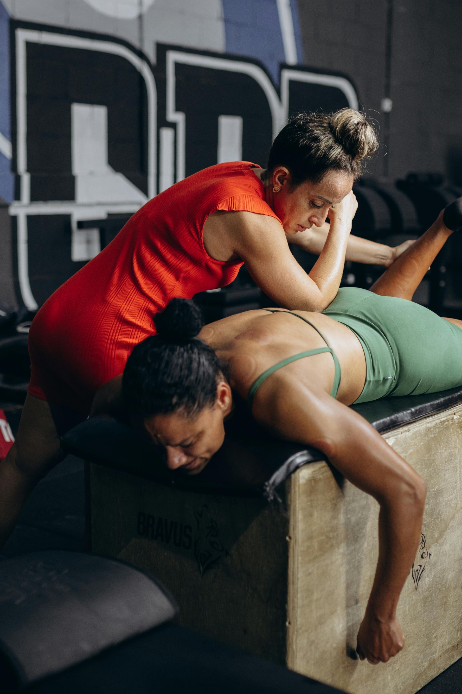 Woman in red shirt massaging another woman's back in a gym setting.