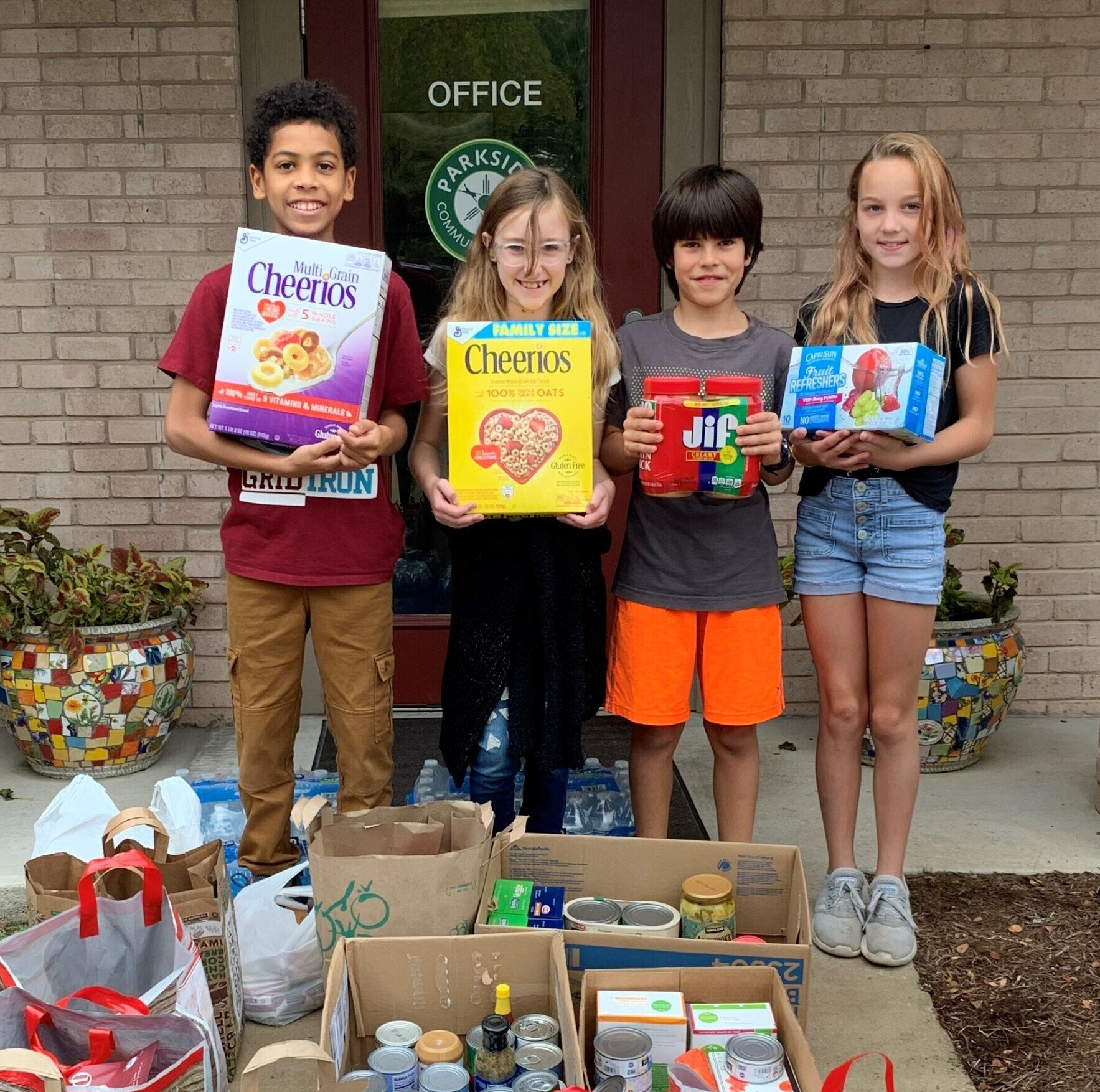 A group of children standing in front of an office holding boxes of food for community service