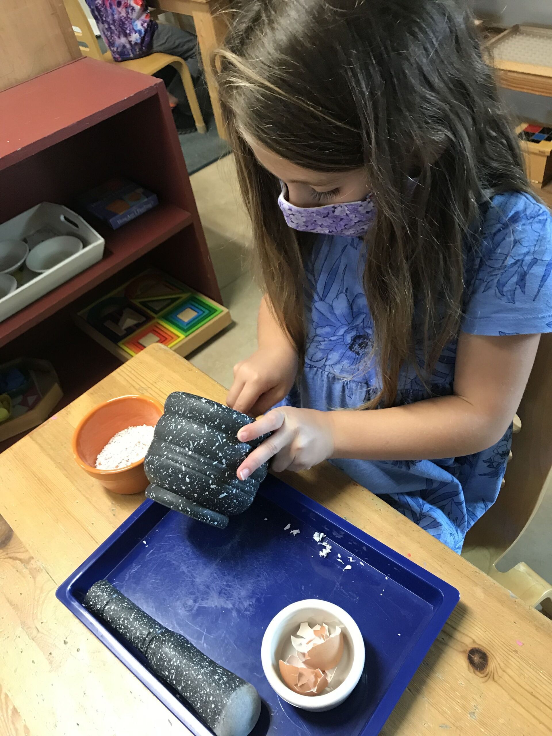 A little girl wearing a mask is sitting at a table cooking in a montessori class
