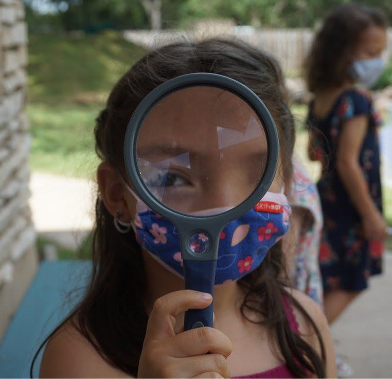 A little girl wearing a mask is looking through a magnifying glass
