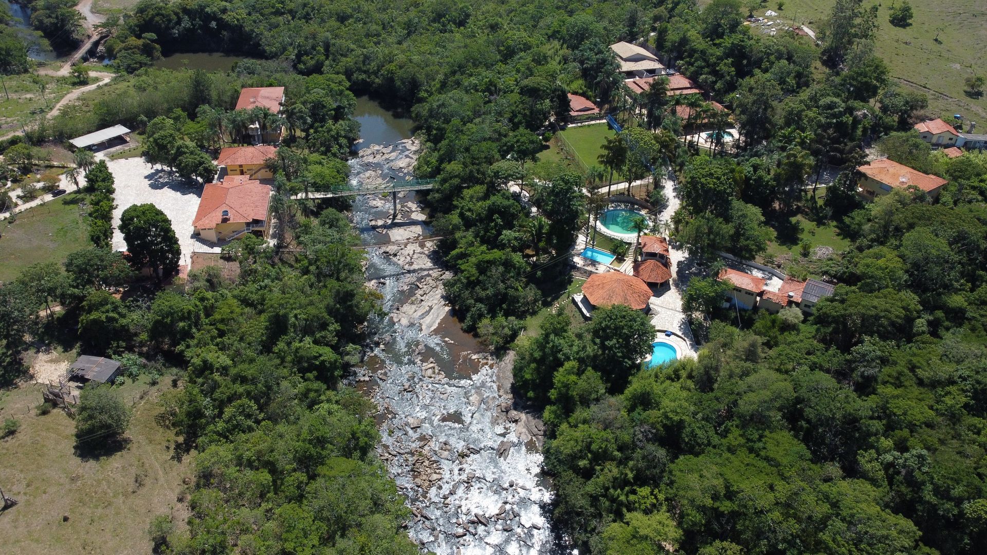 Vista aérea de um rio rochoso que atravessa uma vegetação densa e casas com telhados vermelhos e azuis.