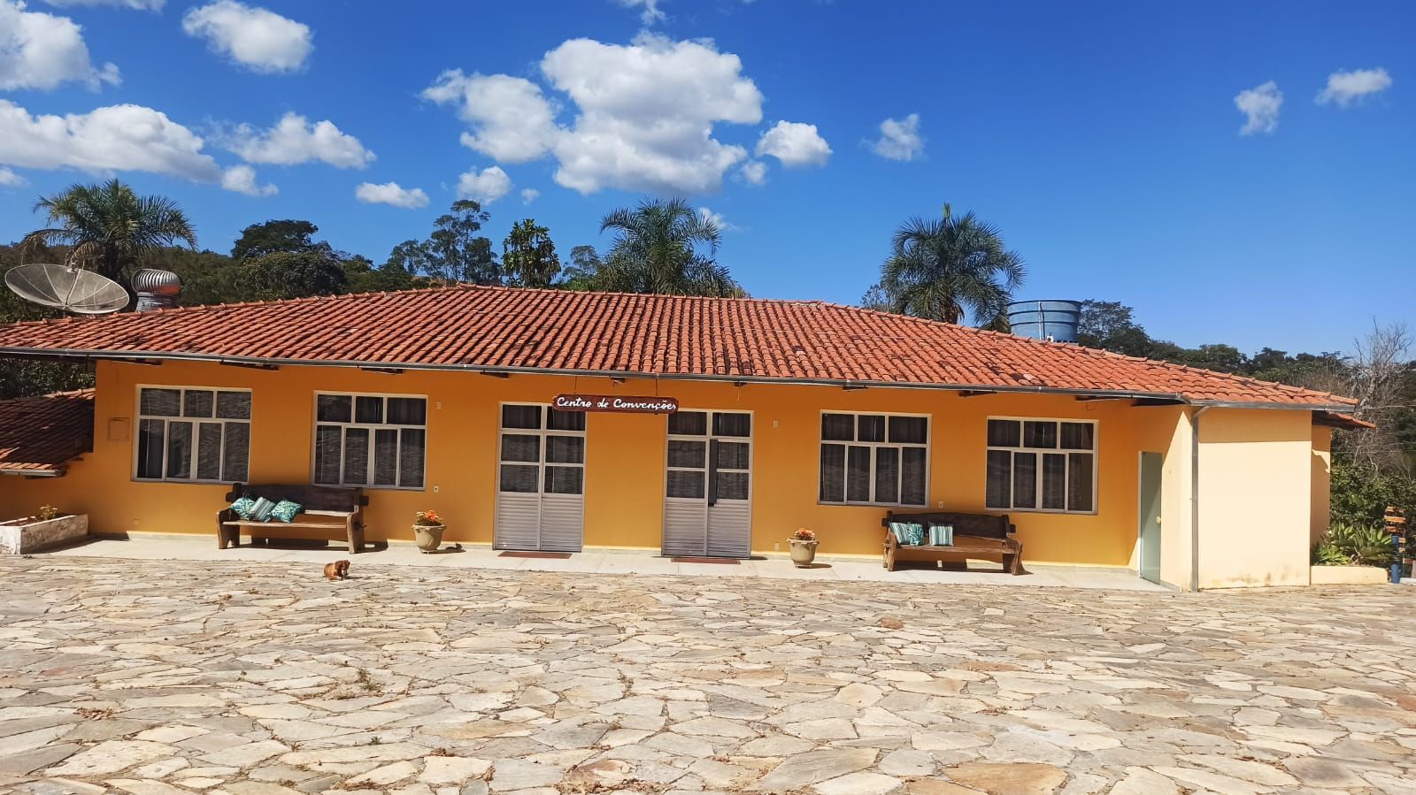 Prédio amarelo com telhado de telha vermelha, janelas frontais e bancos em um pátio de pedra.