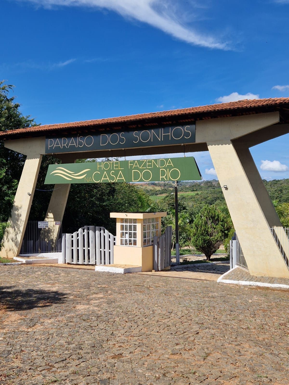 Portão de entrada do Parque Ecológico São João sob um céu azul, com um pequeno quiosque e árvores nas proximidades.