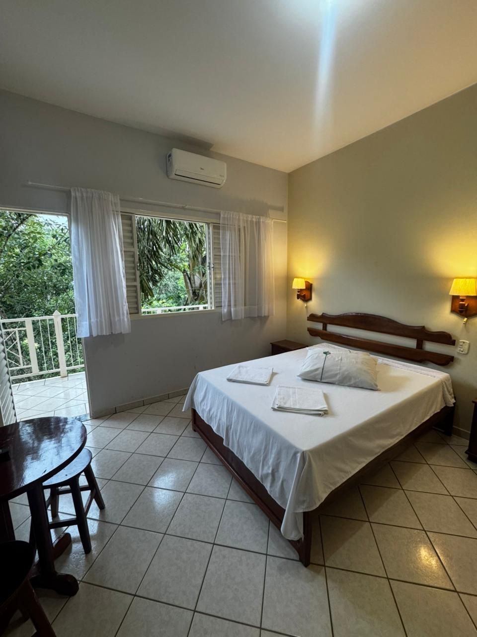 Quarto luminoso com cama de casal, piso de azulejo, janelas amplas e varanda com vista noturna.