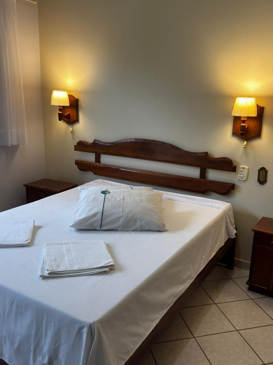 Quarto de hotel impecável com cama de casal, lençóis brancos, apliques de parede e piso de azulejo.
