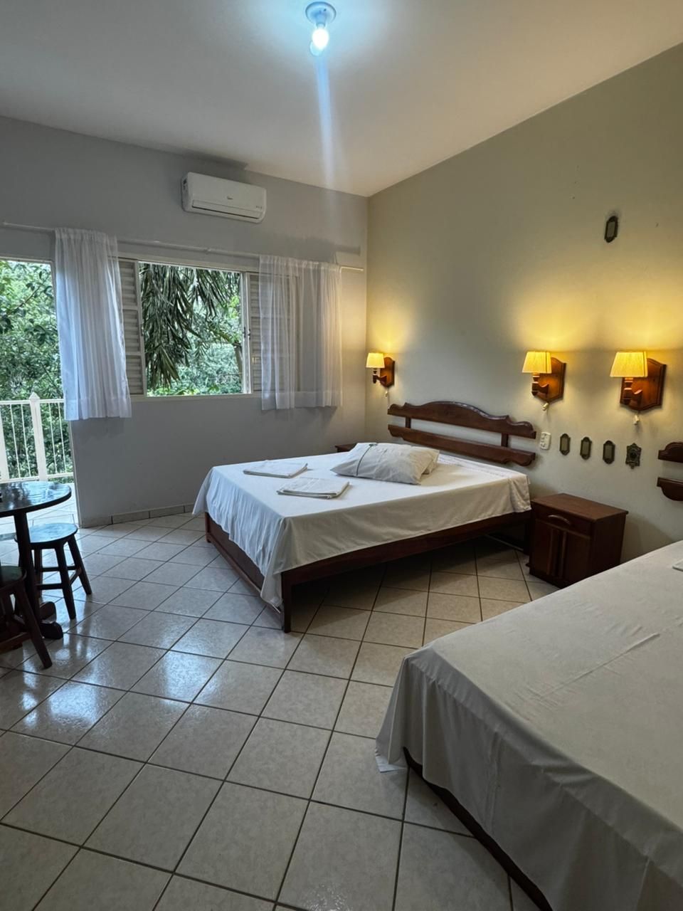 Quarto de hotel luminoso com duas camas, piso de azulejo, candeeiros de cabeceira e porta de varanda com cortinas.