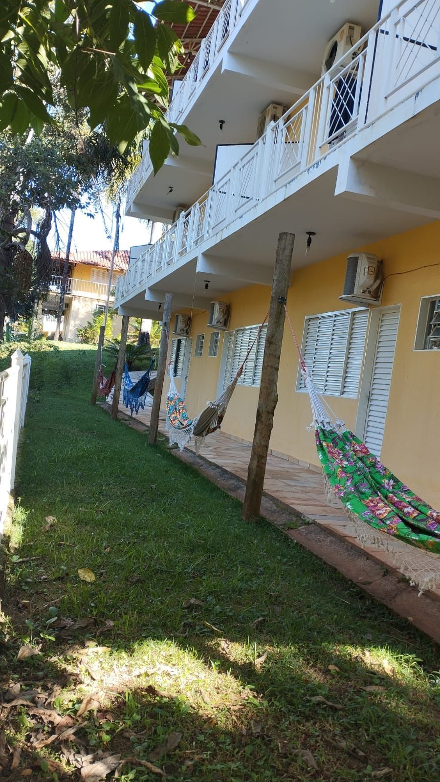 Pátio ao lado de um prédio de apartamentos amarelo com varandas brancas e redes coloridas à sombra.