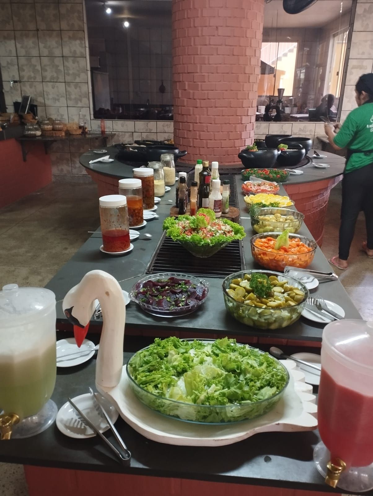 Mesa de buffet com saladas, bebidas e condimentos em uma área de refeições de restaurante.