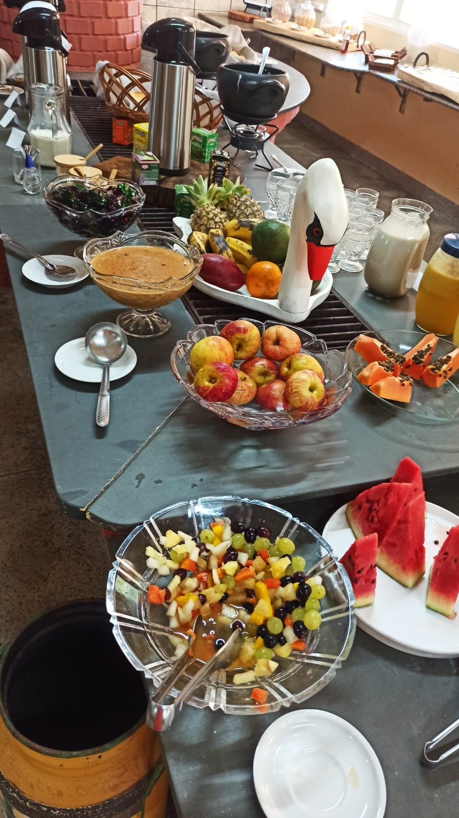 Mesa de buffet com travessas de frutas, doces, bebidas e sobremesas sobre uma toalha de mesa cinza.