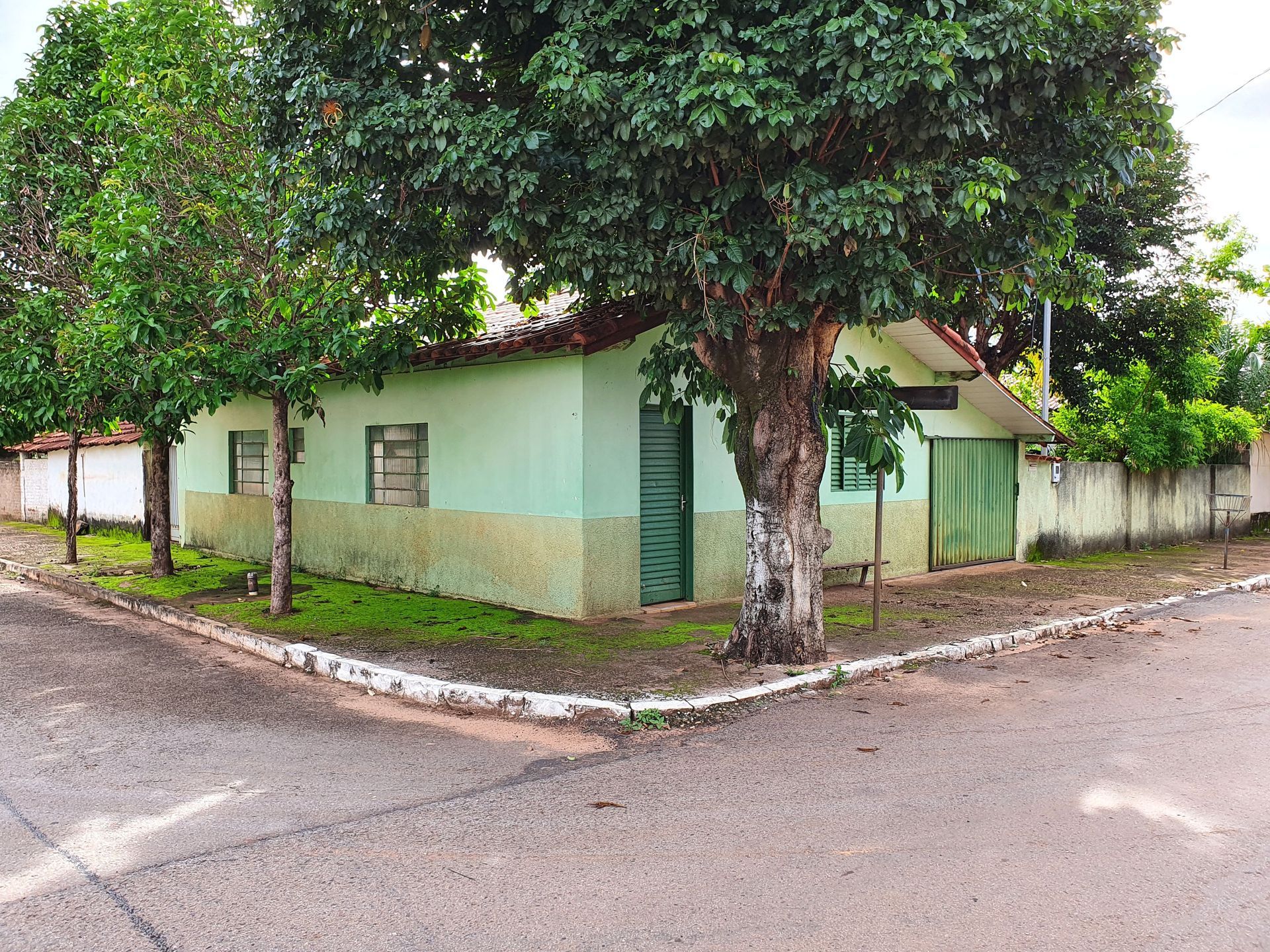 Casa de esquina com paredes verde-claras e telhado vermelho ao lado de uma rua arborizada.