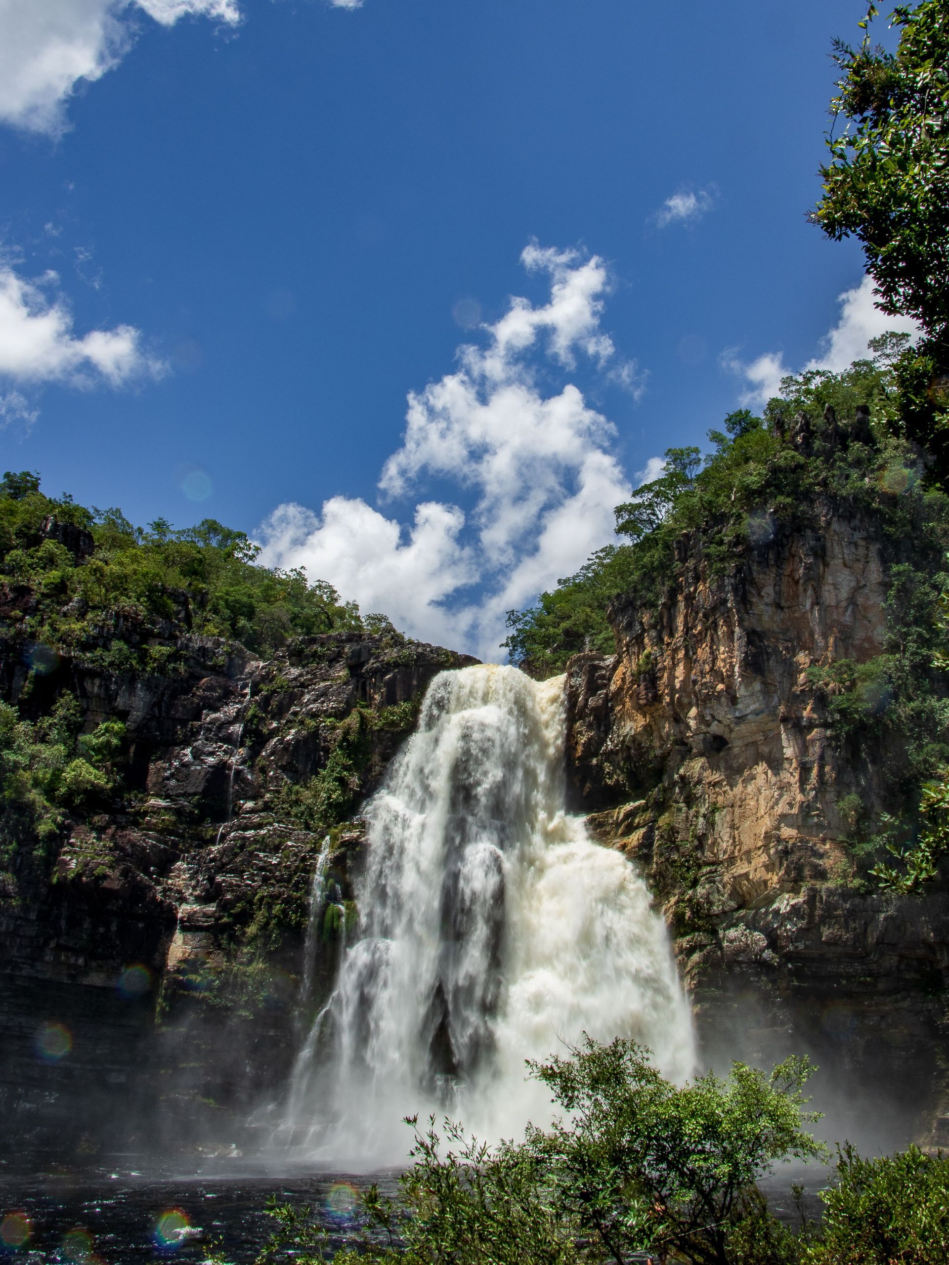 Cachoeira despencando entre penhascos rochosos sob um céu azul brilhante com nuvens.