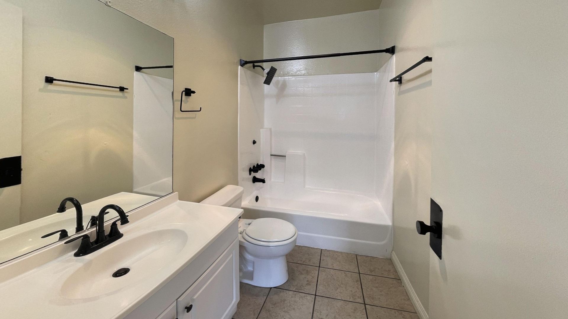 Bathroom with white vanity, toilet, shower, and tan tile floor. Black fixtures and mirror.