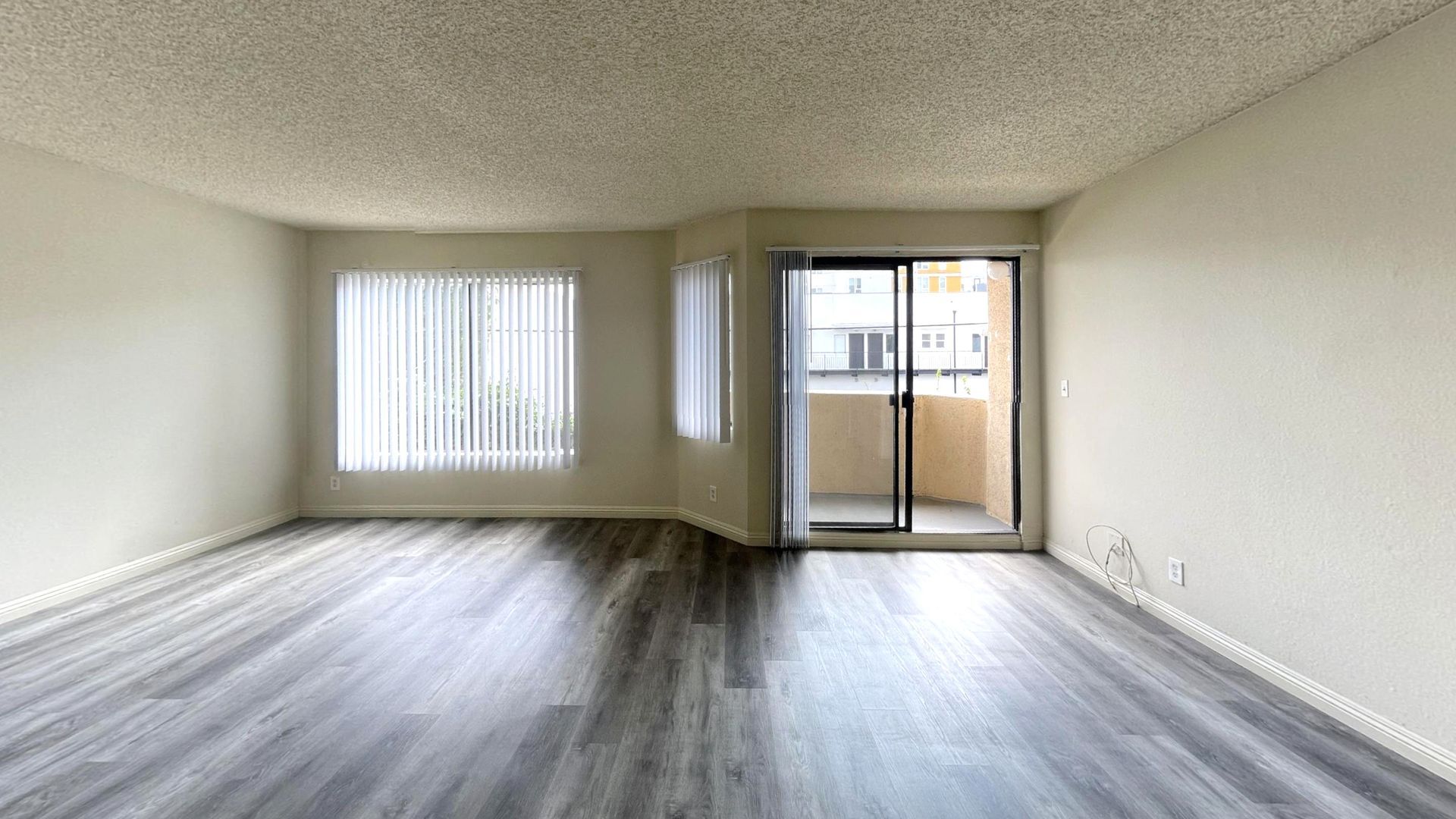 Empty living room with wood-look flooring, large window with blinds, and sliding glass door to a balcony.