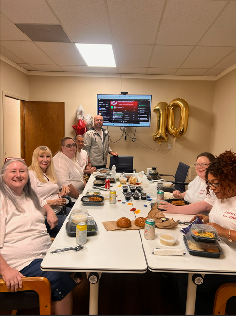 People gathered around a long table at a birthday celebration with gold “10” balloons and food.