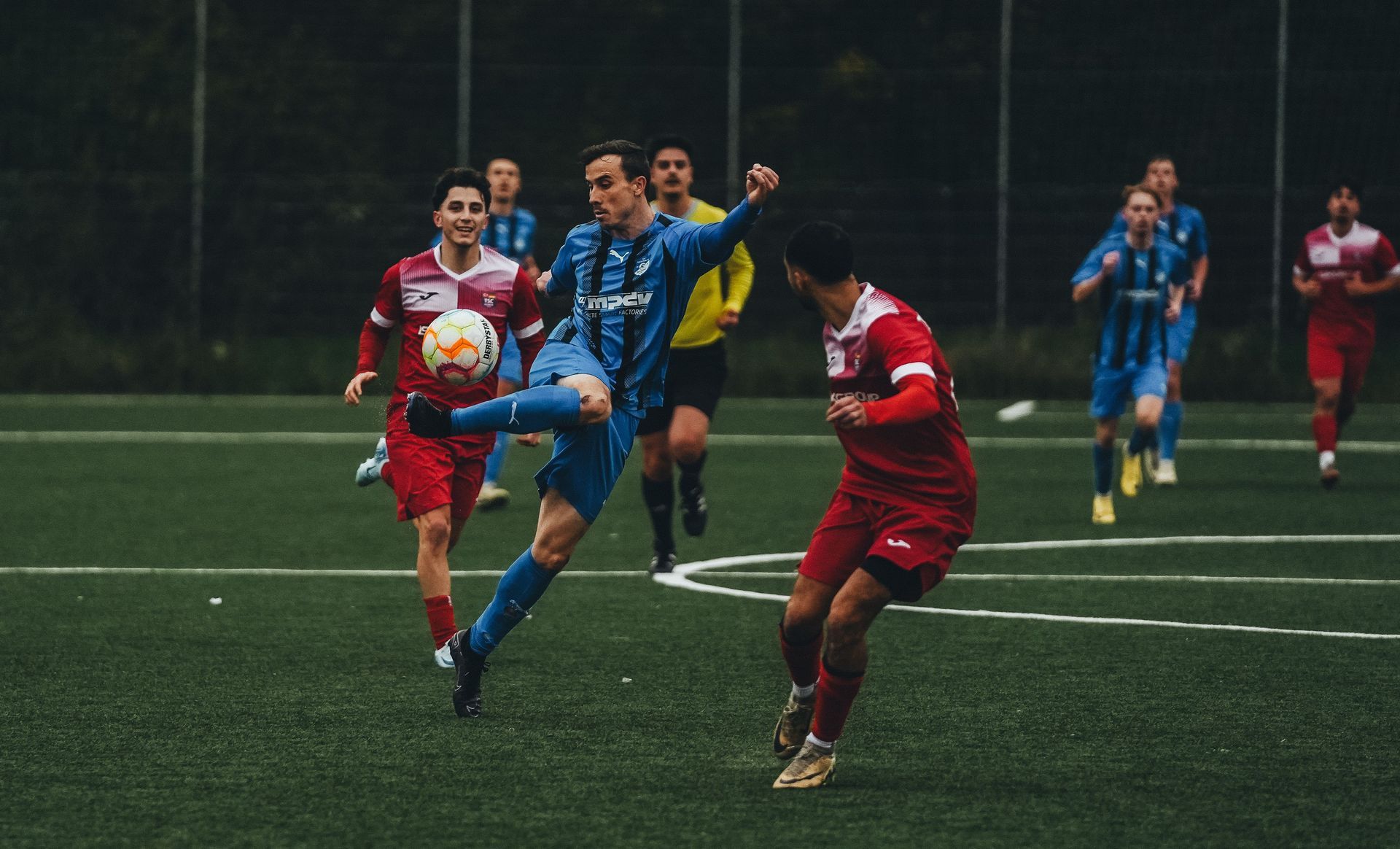 Soccer players in red and blue uniforms contest the ball on a wet field.
