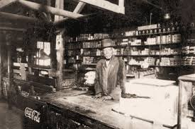Man in hat behind counter in old store, shelves with goods behind him, Coca-Cola sign.