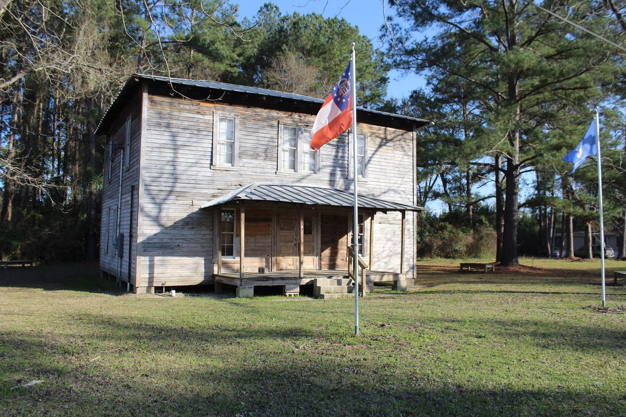 Two-story weathered wooden building with flags in a grassy area, trees in the background.
