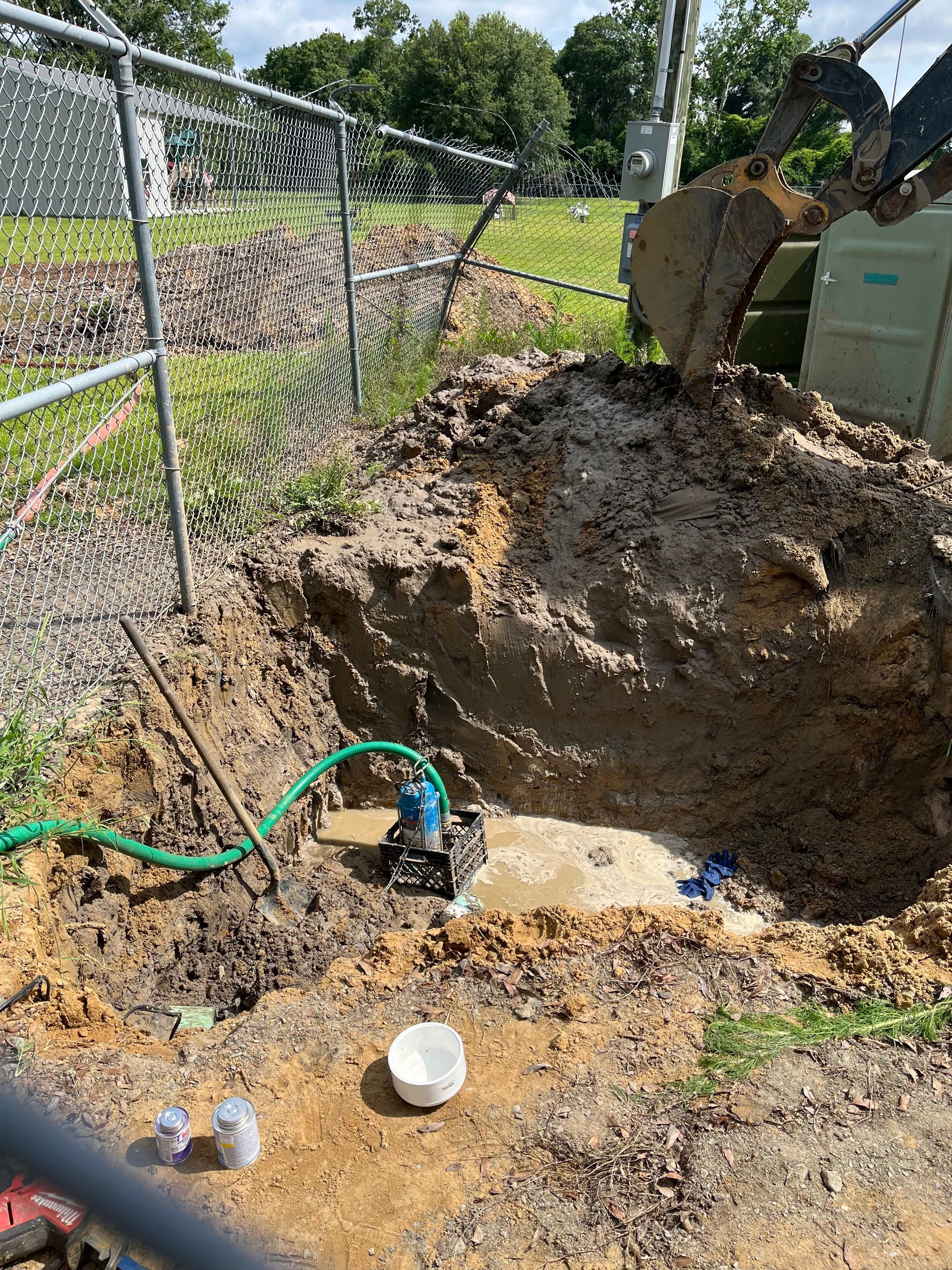 Excavator digging a hole near a chain-link fence, a pump with a green hose at the bottom of the hole.