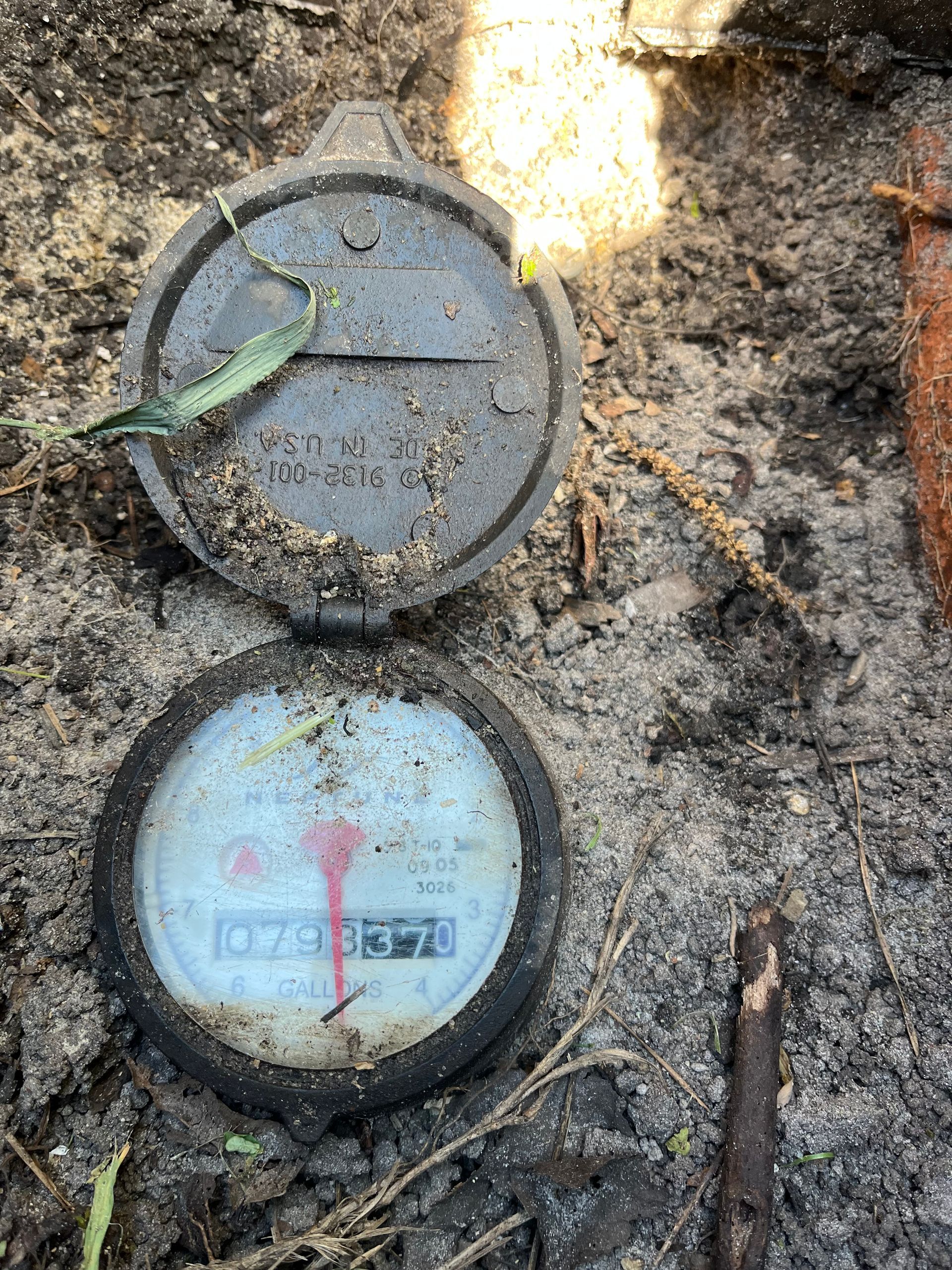 A partially buried water meter with the lid open, displaying numbers 10793678.