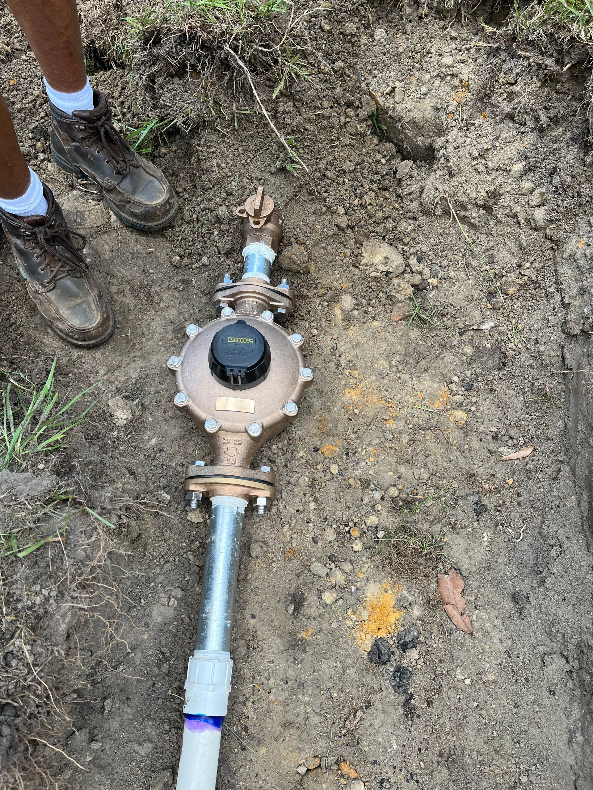 Water meter installed in a ditch with dirt and grass nearby. Person's muddy boots visible.