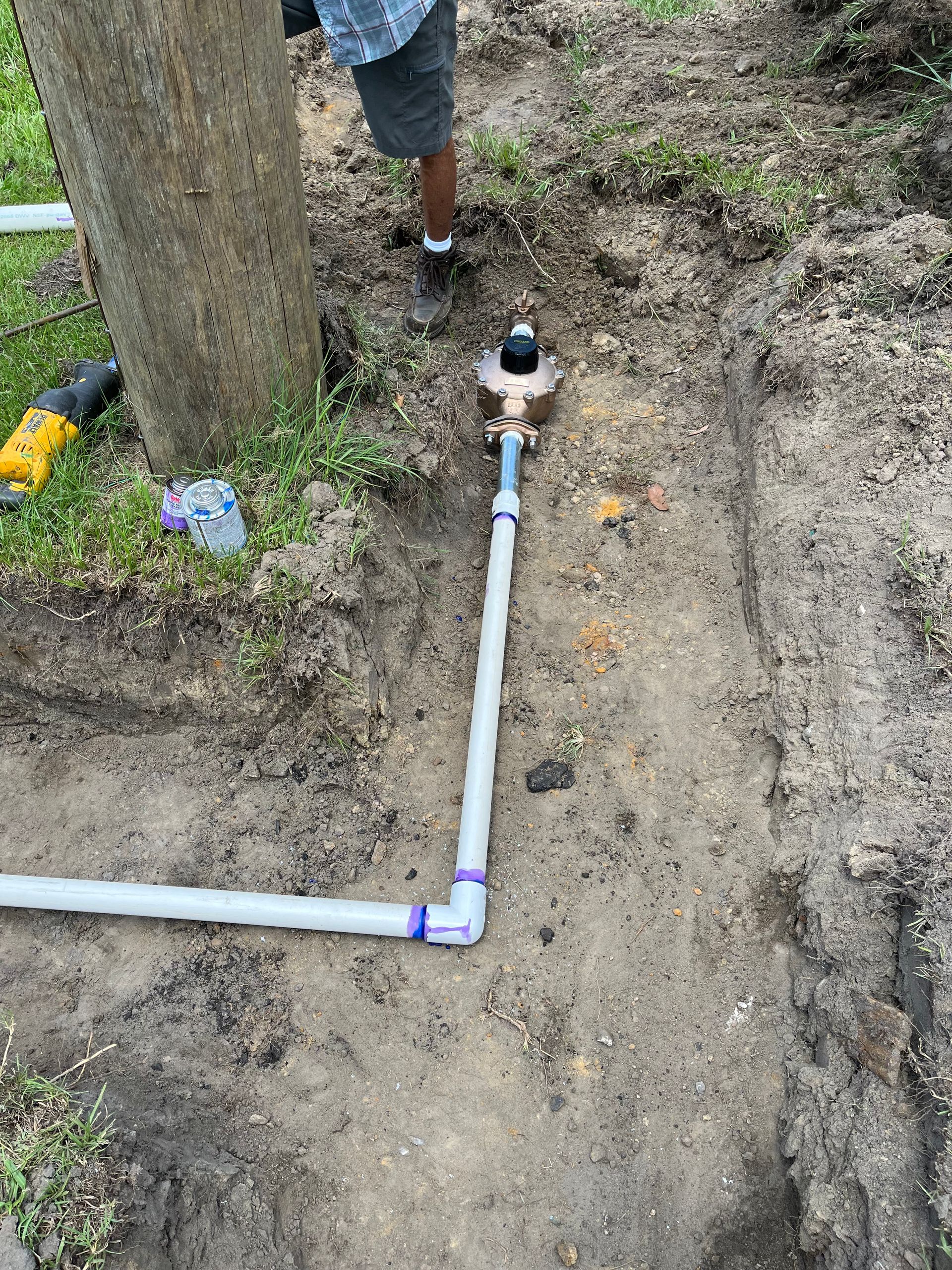 Pipes being installed in a trench. A person stands nearby. Tools and a tree are also present.