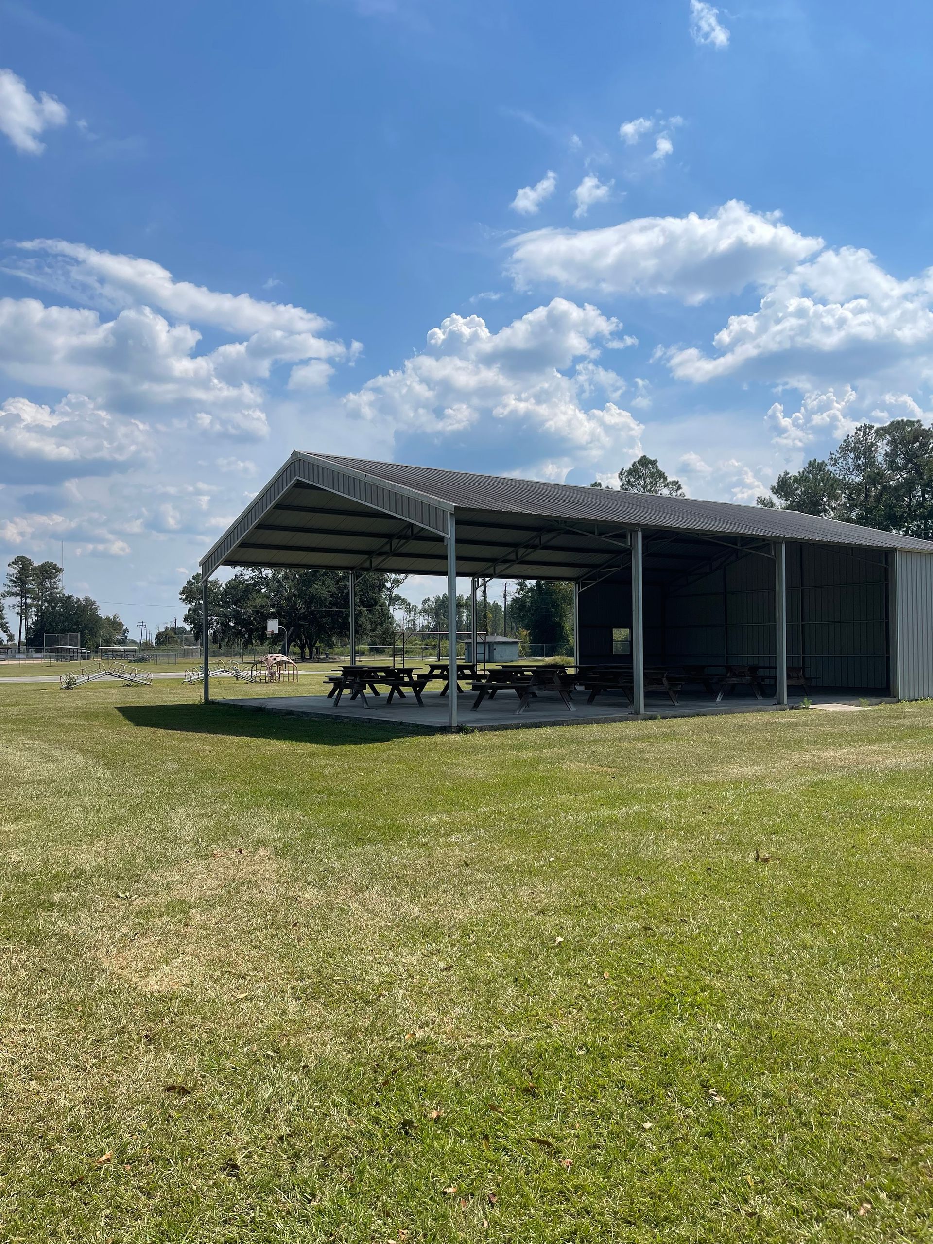 A picnic shelter with picnic tables in a grassy field on a sunny day with clouds.