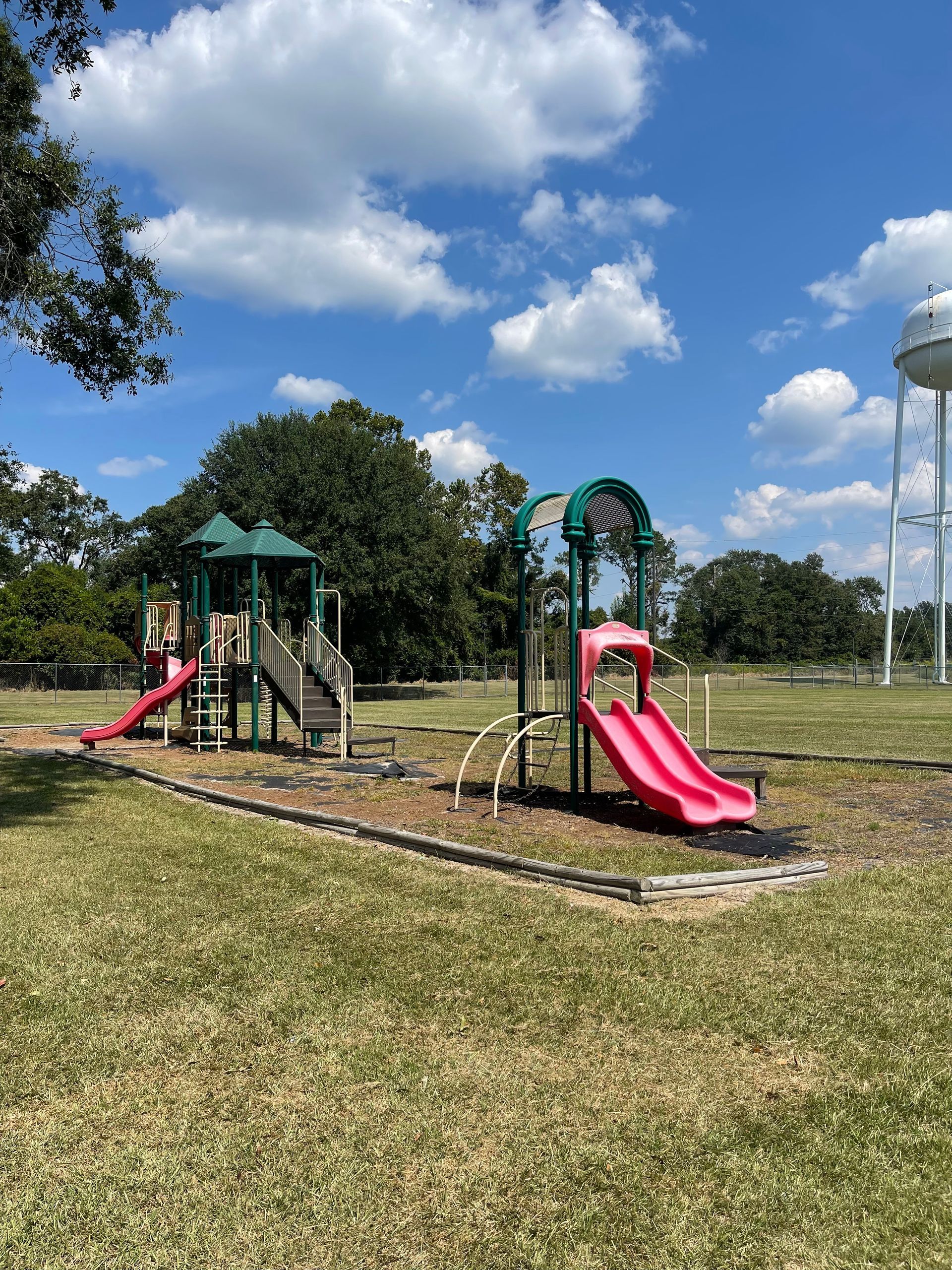 Playground with two slides, stairs, and a climbing arch on a grassy field; water tower in the background under a blue sky.