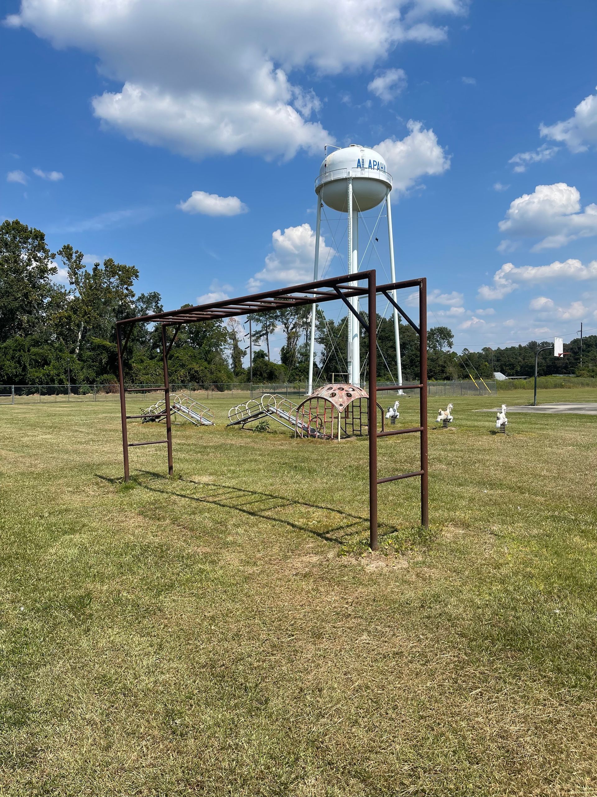 A rusty metal jungle gym in a grassy field with a water tower and blue sky.