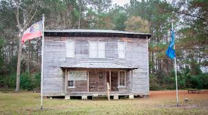 Old, weathered two-story building with porch; Confederate flag and blue flag flank it.  Wooded area in background.