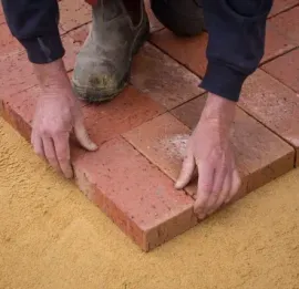 Person kneeling, laying red bricks in a herringbone pattern on a bed of sand.
