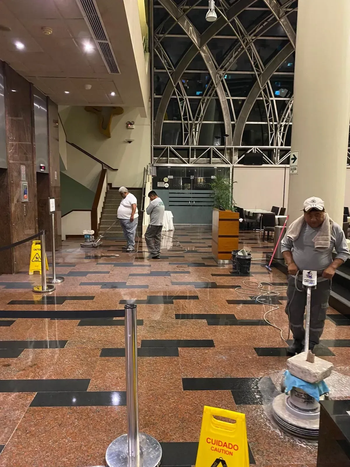 Workers cleaning a large, multi-colored tiled floor in an indoor lobby. Wet floor warning signs are present.