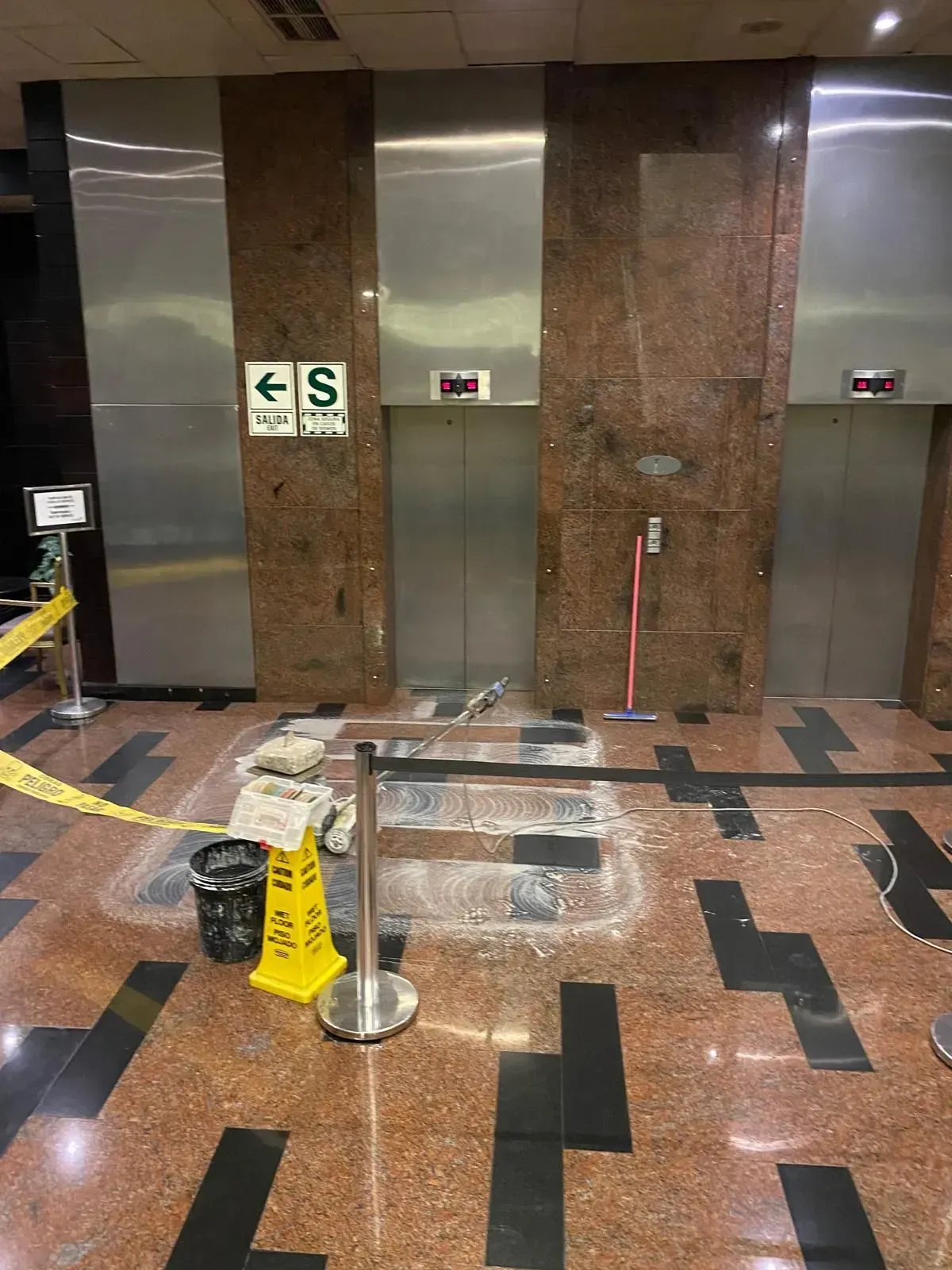 Elevator lobby with damaged floor. Caution tape, wet floor sign, and debris near elevators.