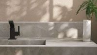 Bathroom vanity with matte black faucet, gray countertop, white vase with greenery, and beige textured wall.