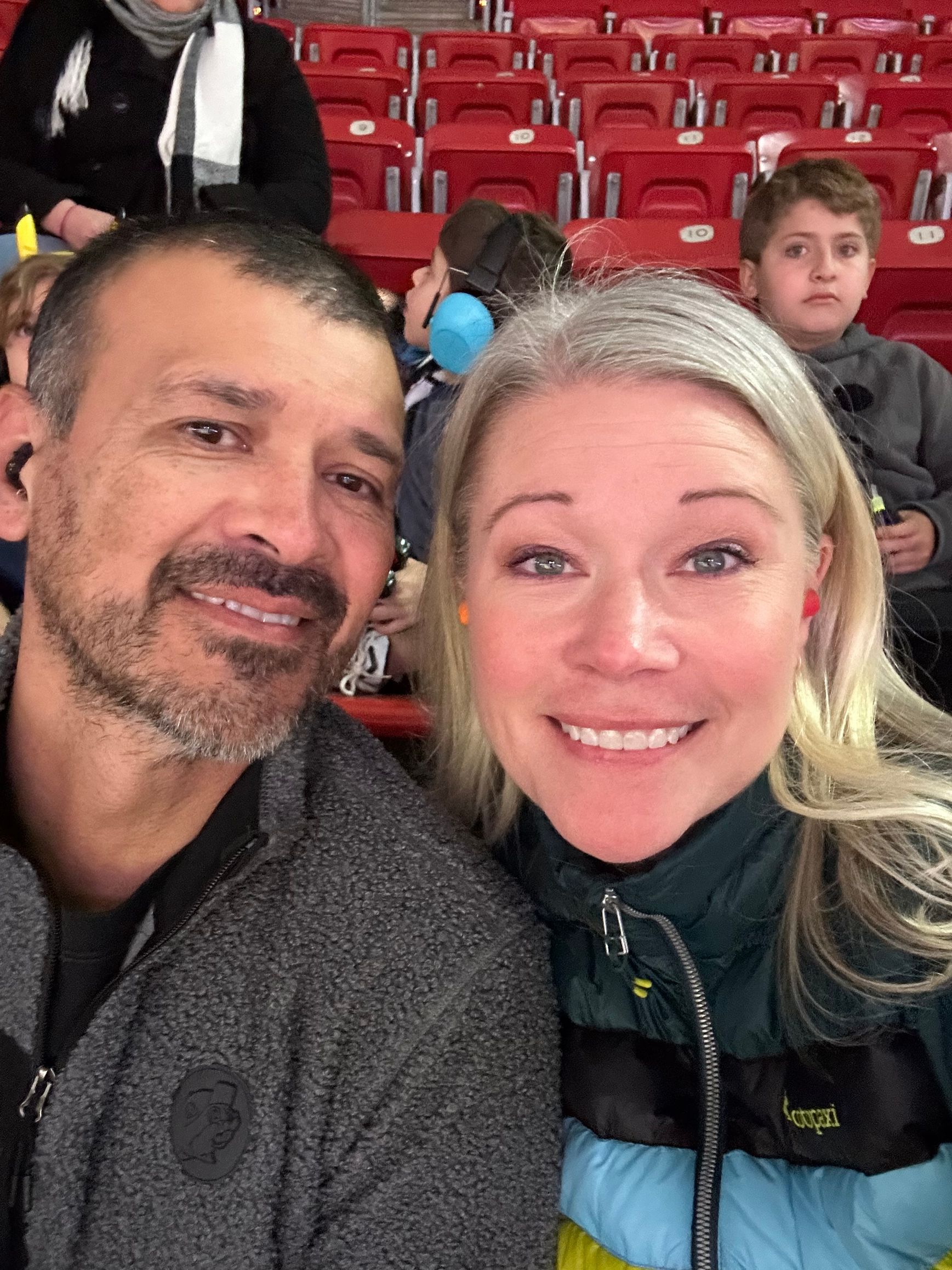 Two smiling fans taking a selfie in red stadium seats, with a child in the background.