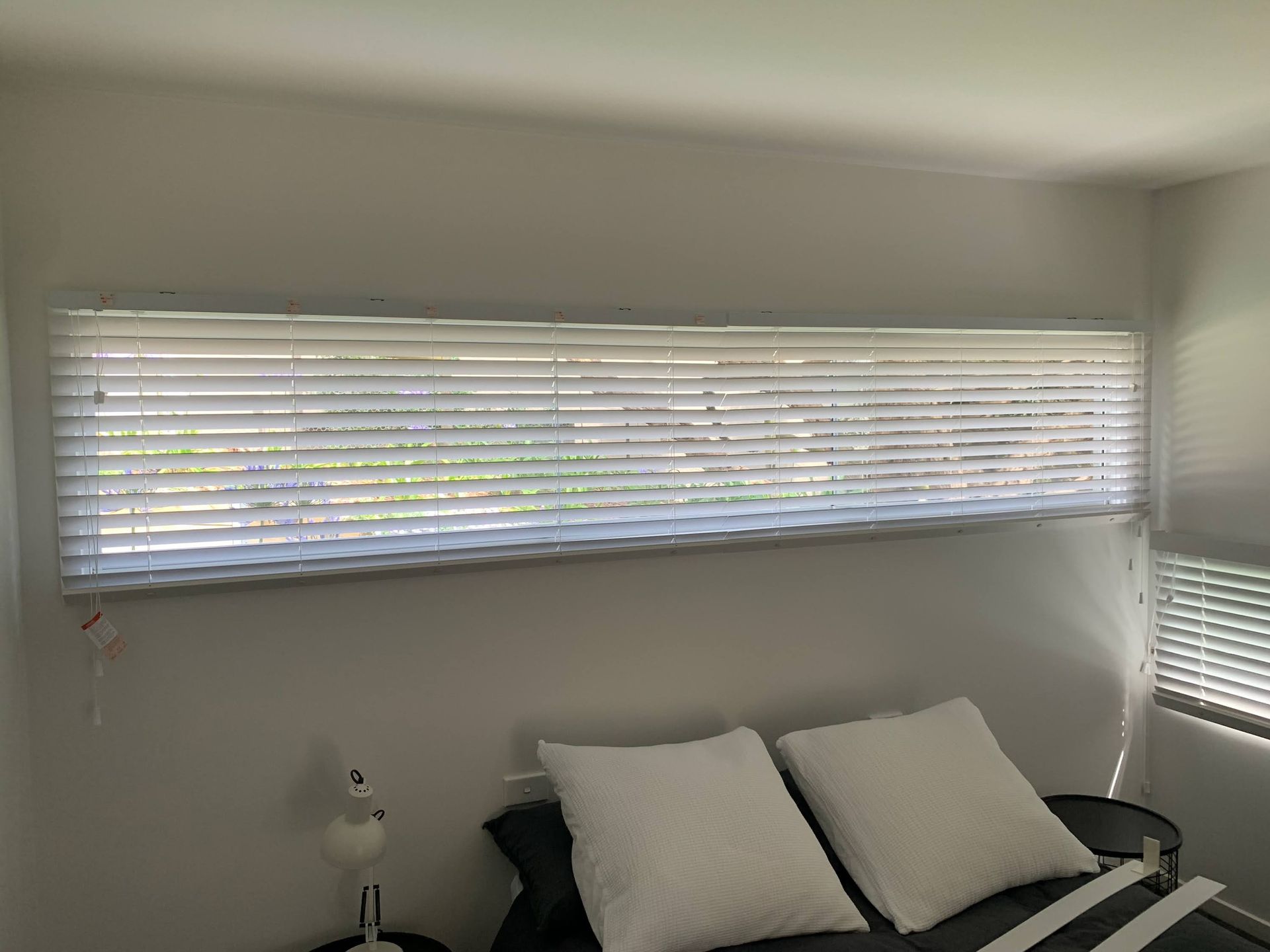 Small room with a window, white blinds, and a couch with white pillows beneath it.