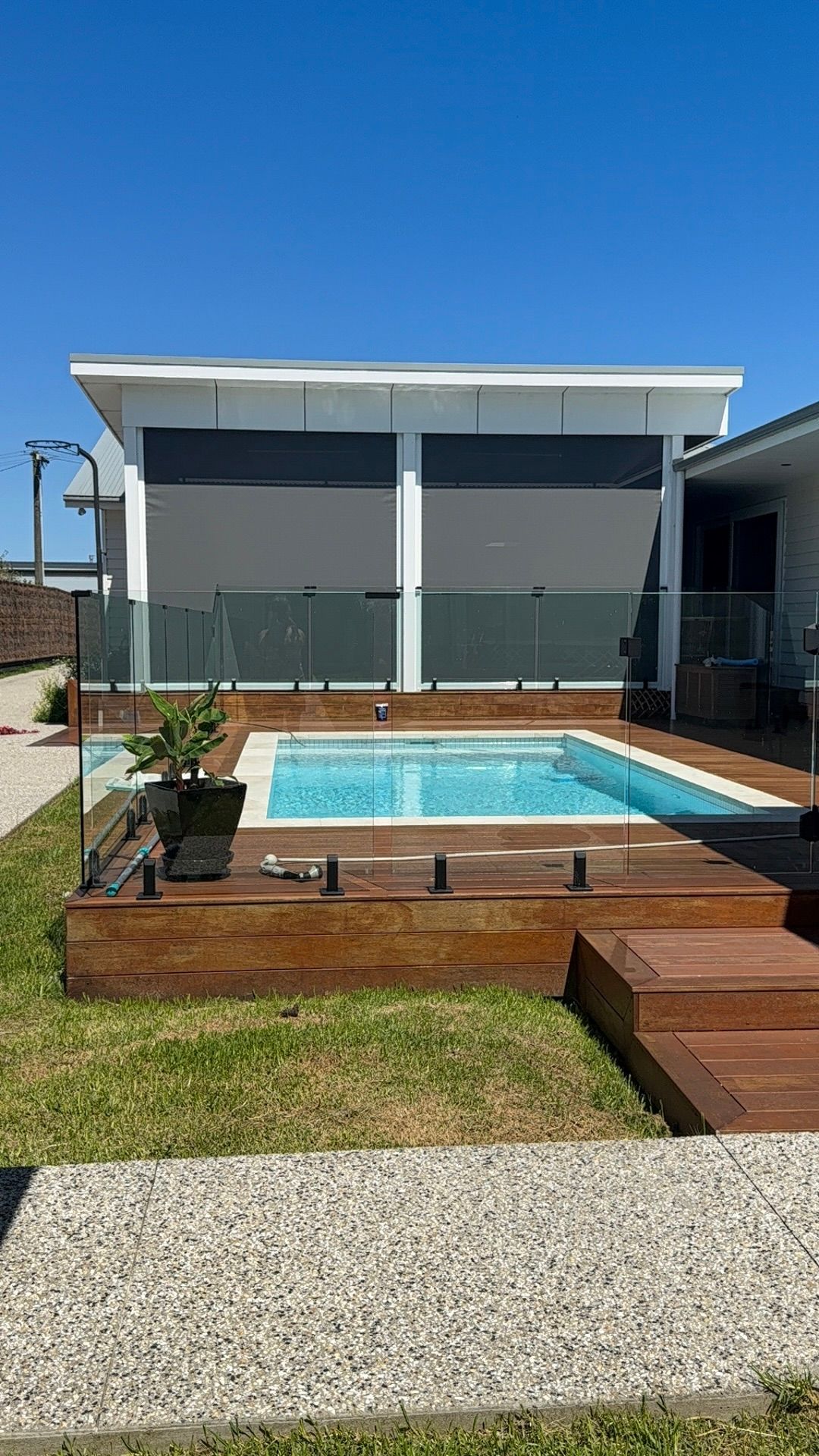 Modern backyard pool with wooden deck, steps, potted plant, and shade sail under a blue sky