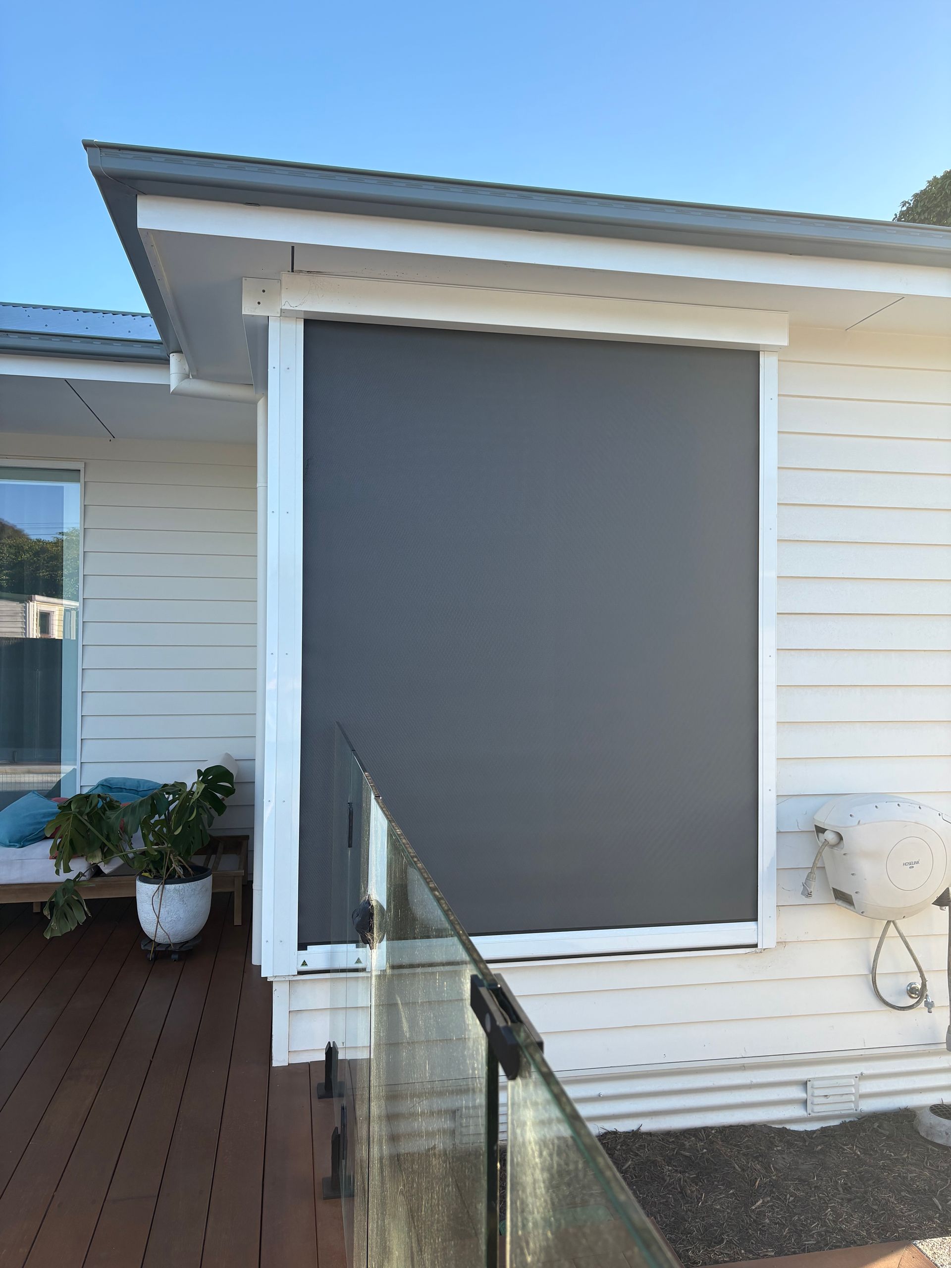 Patio with a large gray roller blind on a white house exterior, viewed from a wooden deck and railing
