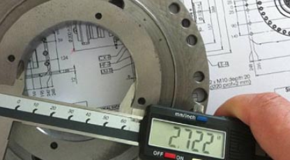 A digital caliper measuring a metal machine part resting on an engineering blueprint.