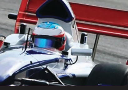 Formula race car driver in a white and blue car, close-up on the track with red barriers behind.