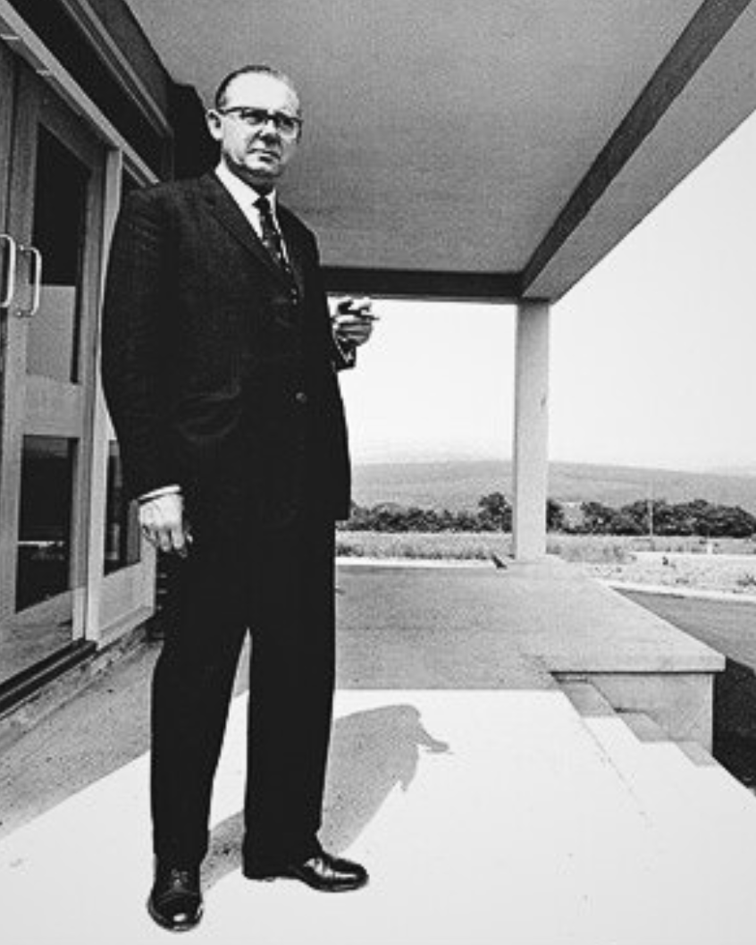 A man in a suit and glasses stands on a building porch, holding a small object, with a landscape in the background.