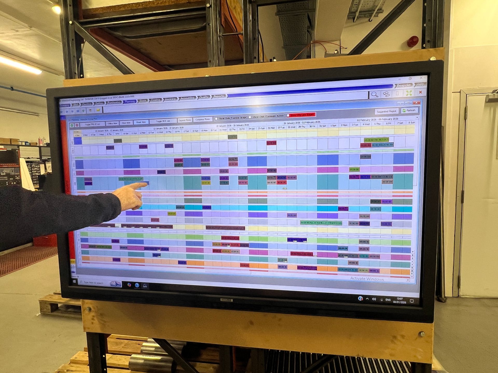 A person points to a large digital screen displaying a colorful project management or production schedule in a warehouse.