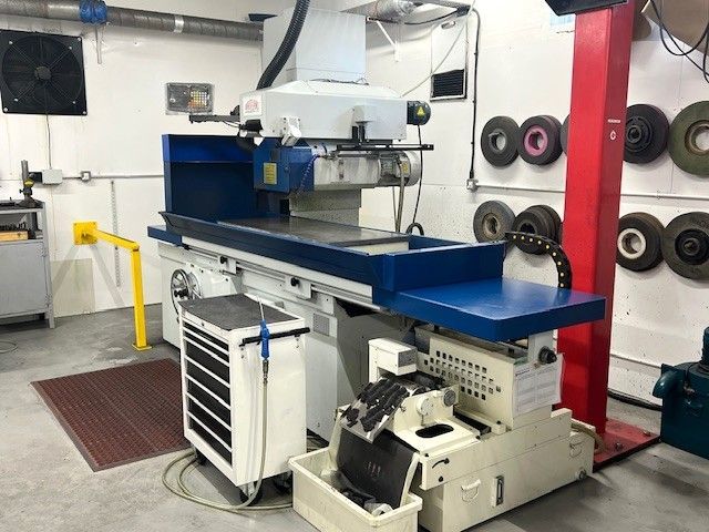 A surface grinder machine sits in a workshop with several grinding wheels mounted on the wall.