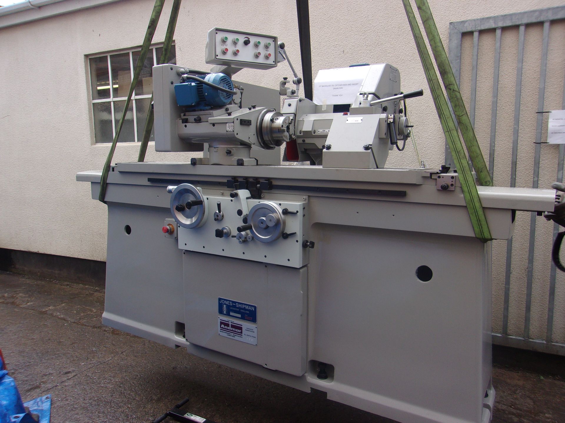 A large, grey industrial cylindrical grinding machine suspended in the air by green lifting straps outdoors.