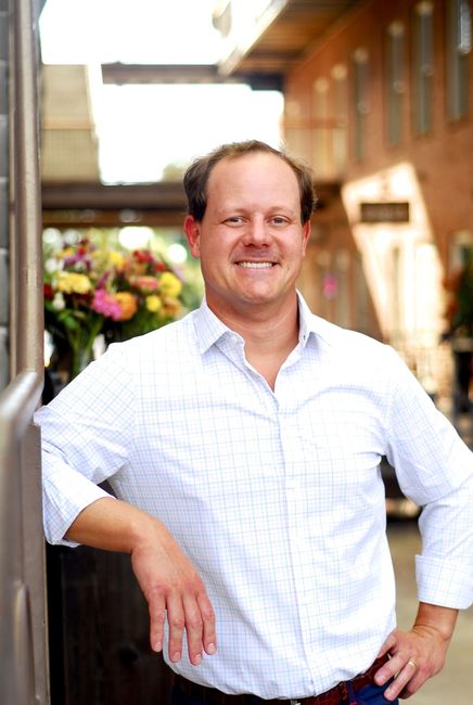 Man in light blue checkered shirt smiling, leaning against a building.