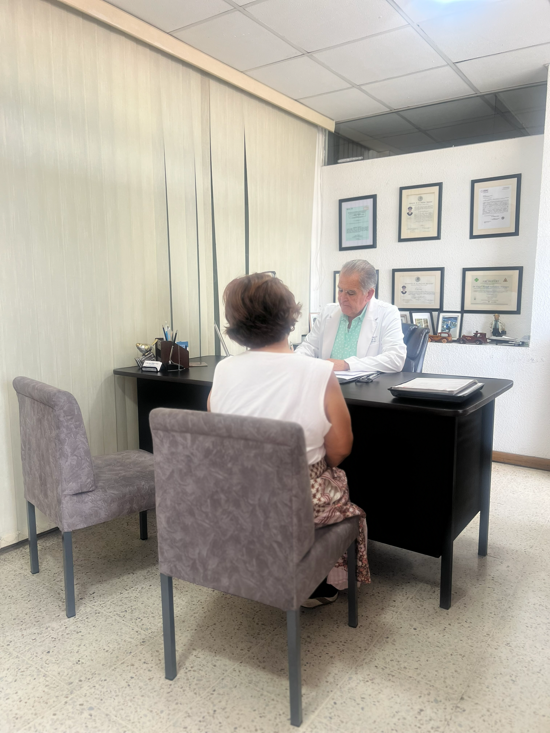 Dos personas conversan frente a frente en una pequeña oficina con certificados enmarcados en la pared.