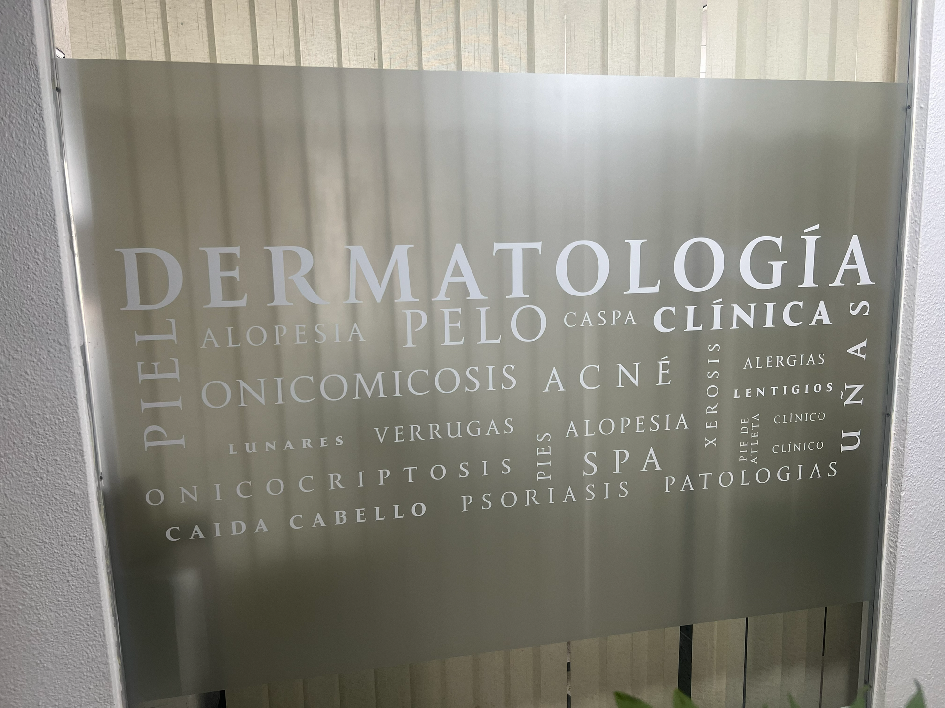 Letrero de cristal para clínica de dermatología y cosmetología, con letras esmeriladas sobre un fondo de cortina blanca.